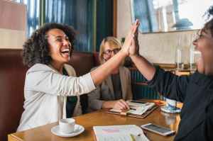 two smiling women sit around a table. One is high fiving an unseen third person. One of the women is black the other is white and wearing glasses